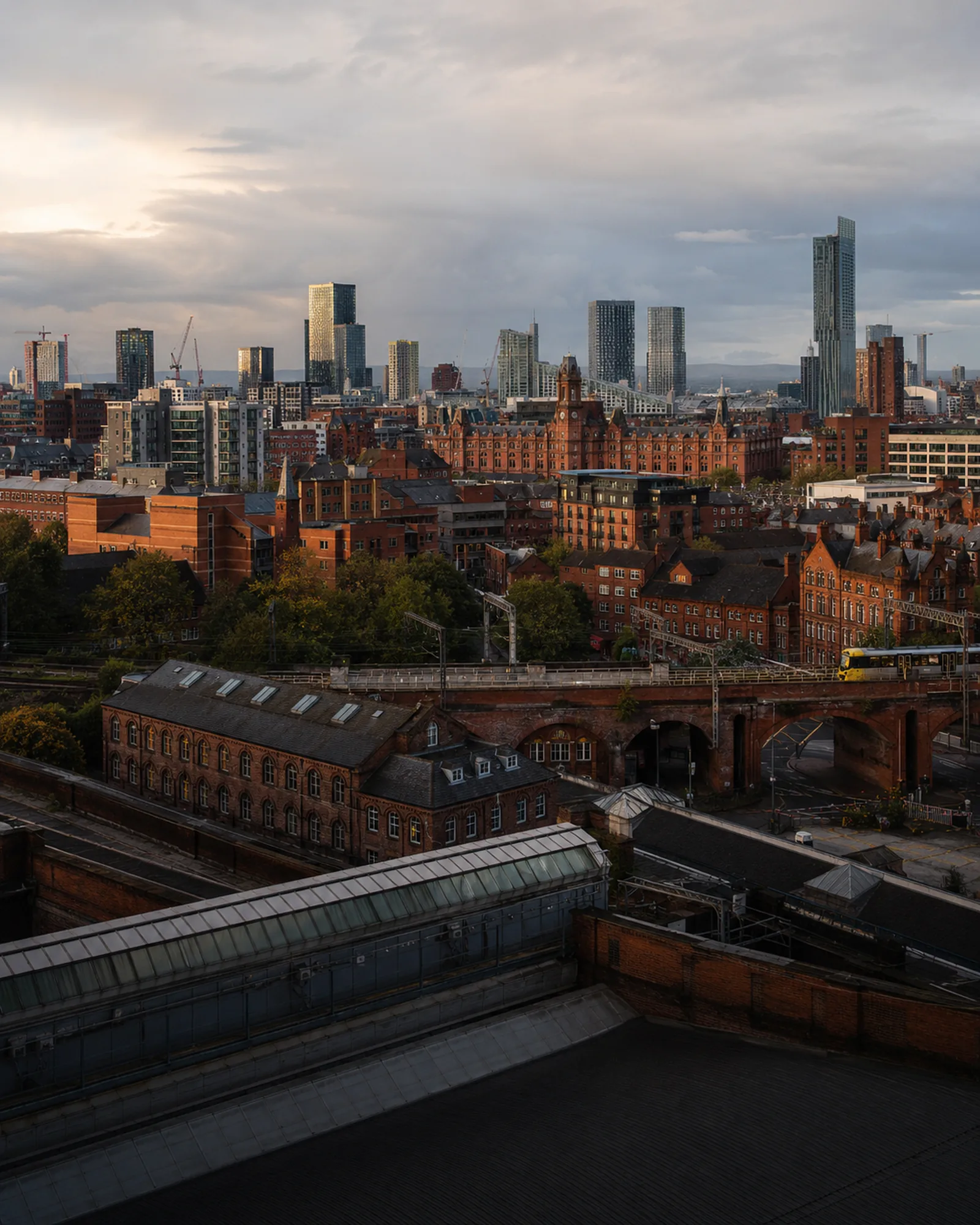 Manchester's Deansgate towers rising behind red-brick warehouses and a railway viaduct at dusk.
